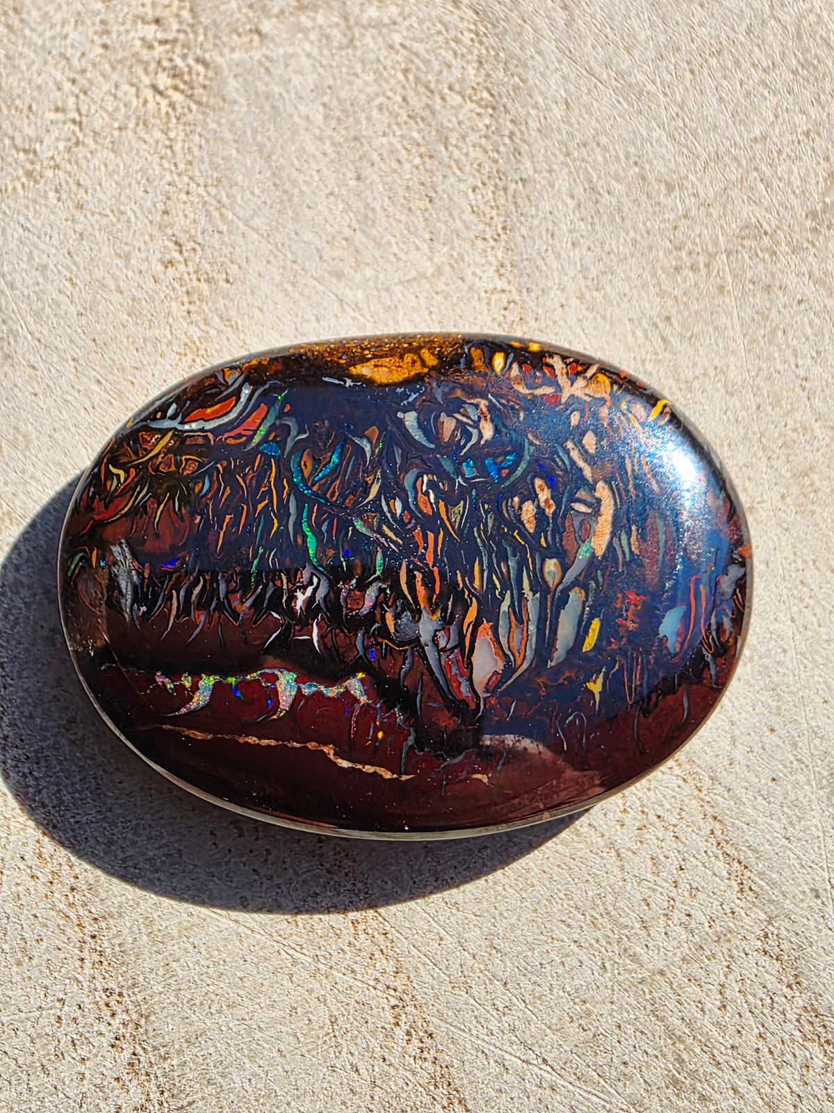 A polished oval boulder gemstone with a highly iridescent, multicoloured play-of-colour pattern over a dark reddish-brown base, resting on a textured beige stone surface.