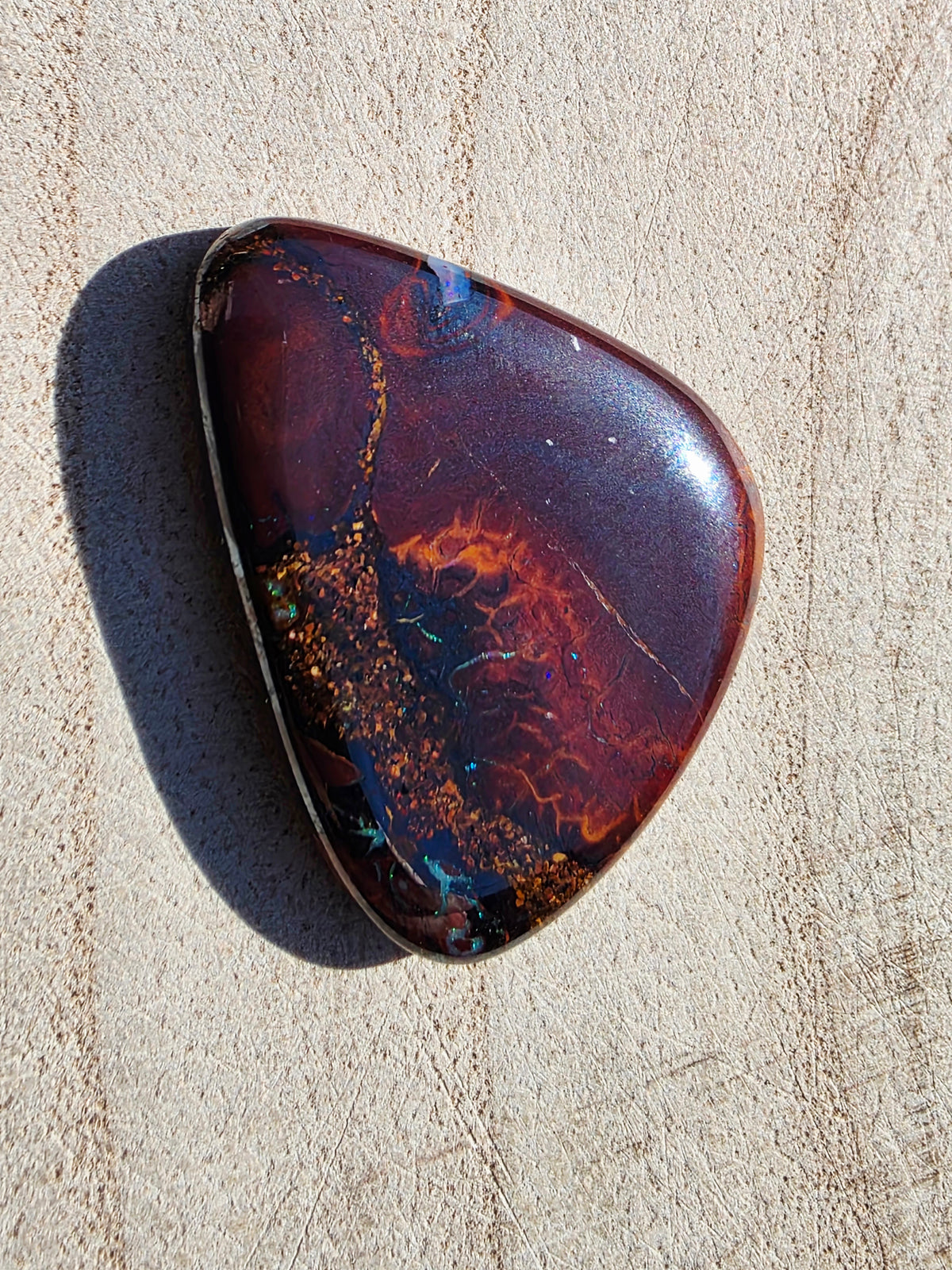 A polished, teardrop-shaped boulder opal with a rich marbled pattern of deep red, purple, orange, and dark blue, resting on a textured beige stone surface.