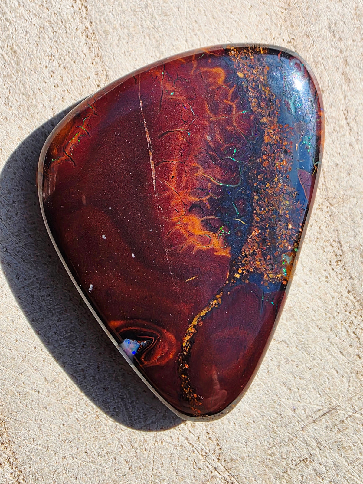 A polished natural opal stone with a teardrop shape, featuring a marbled pattern of deep red, orange, brown, and blue with gold flecks, set in a silver bezel on a light beige textured background.