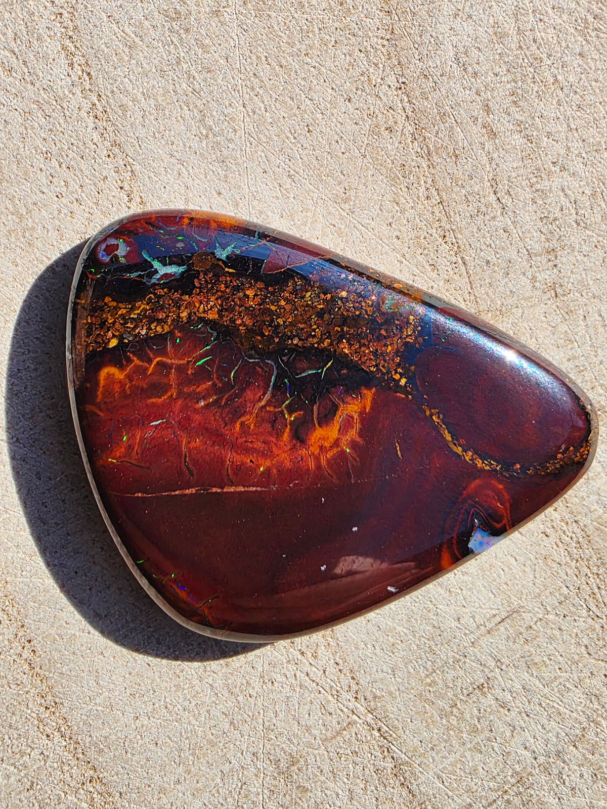 A polished teardrop-shaped boulder opal with rich dark red, orange, and black marbled patterns, resting on a textured beige stone surface.