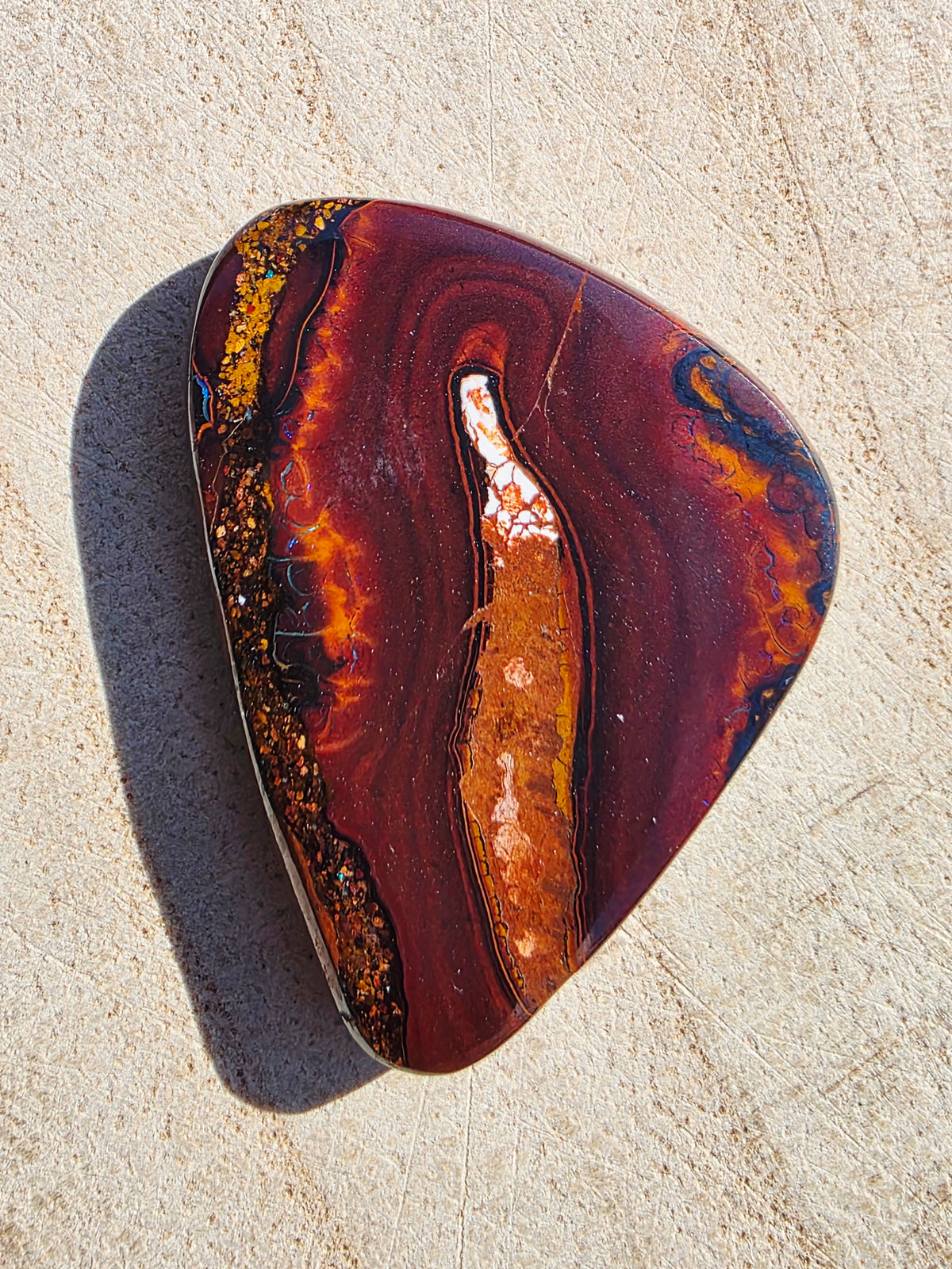 A polished natural stone with swirling bands of deep red, orange, brown, black, and gold, photographed on a light sandy surface with a strong shadow.