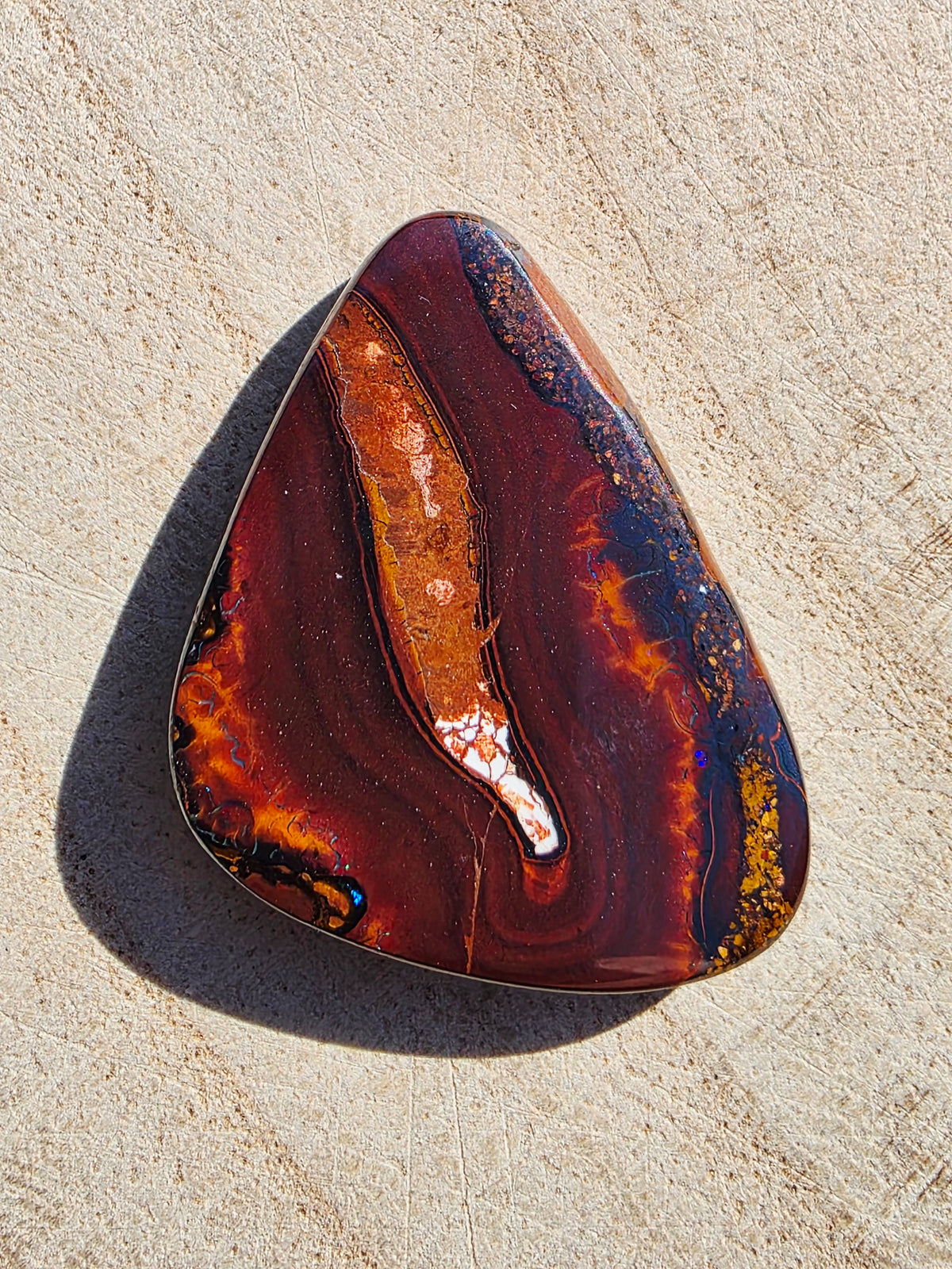 A polished teardrop-shaped opal with swirling bands of dark red, orange, brown, and blue, resting on a textured beige stone surface.
