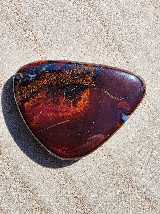 A polished natural opal cabochon with a rich marbled pattern of deep burgundy, orange, gold, and dark blue, set in a silver-toned bezel on a light beige textured surface.