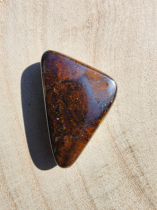 A polished triangular opal stone with iridescent brown, amber, and blue flashes, resting on a light beige textured surface.