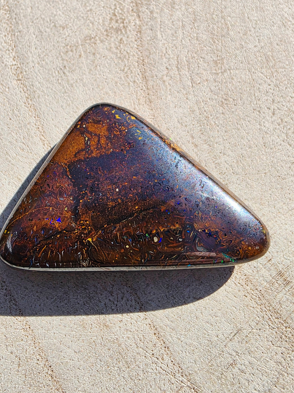 A polished triangular brown opal gemstone with iridescent flecks and a silver bezel, resting on a textured beige stone surface.