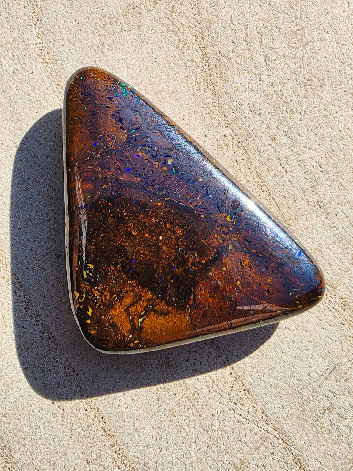 A polished triangular opal gemstone with a rich brown, amber, and black marbled pattern and subtle iridescent flashes, set in a silver bezel and resting on a sandy beige surface.