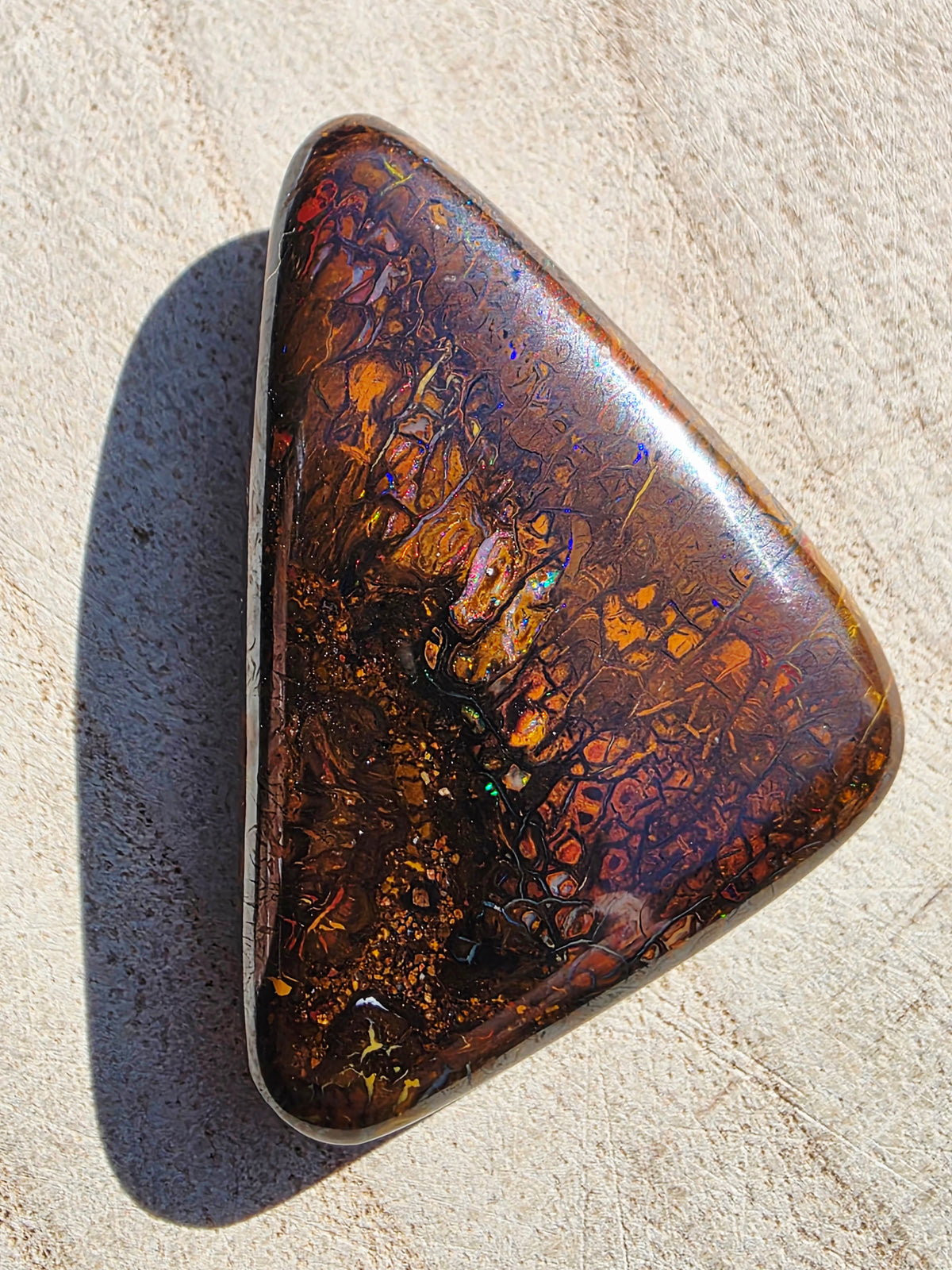 A polished triangular boulder opal gemstone with a rich pattern of brown, gold, and orange iridescence, resting on a light beige textured surface.