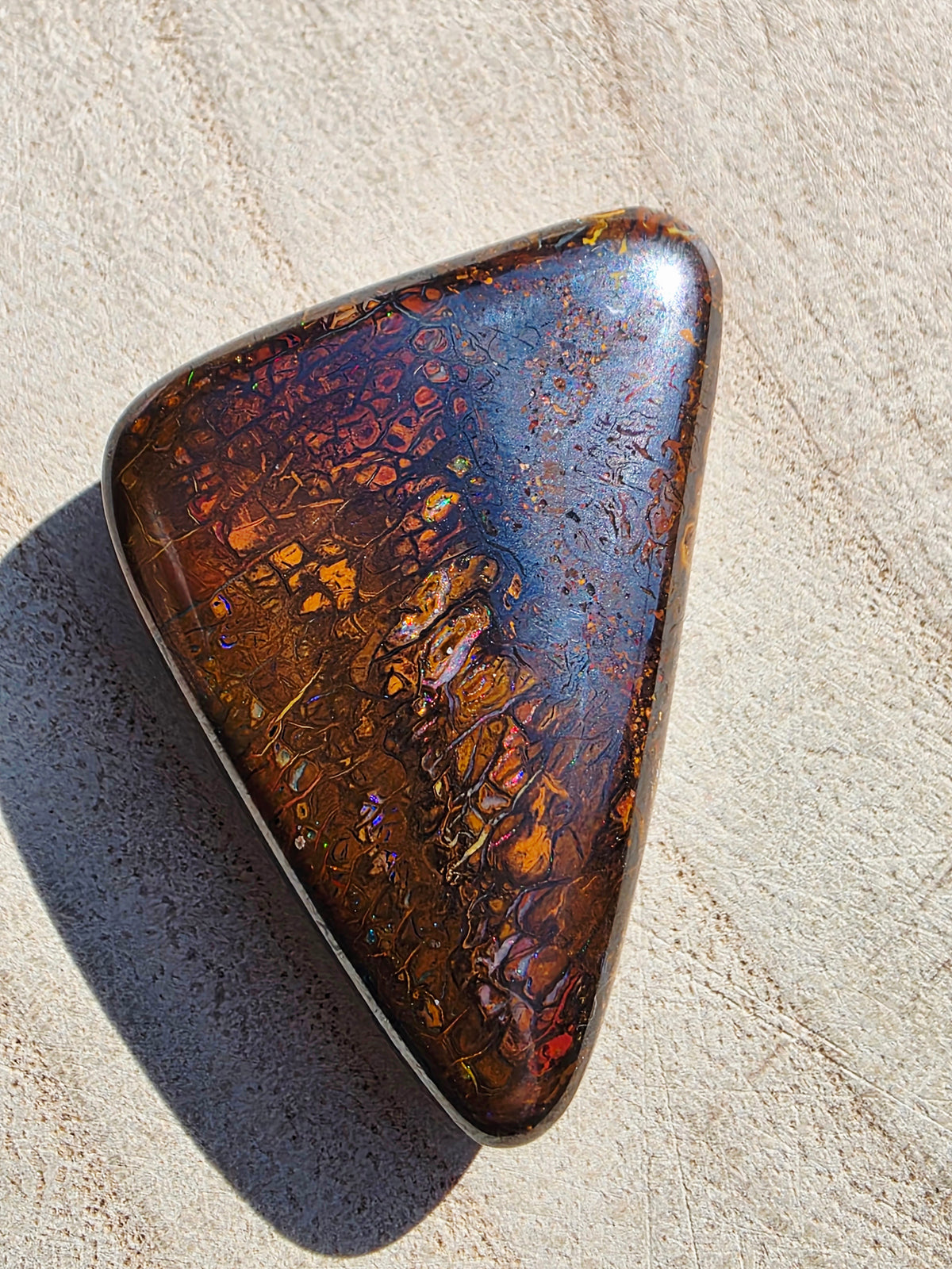 A polished triangular opal stone with a rich iridescent play-of-color, featuring a mix of deep blue, coppery orange, and brown tones, set in a silver bezel on a textured beige surface.