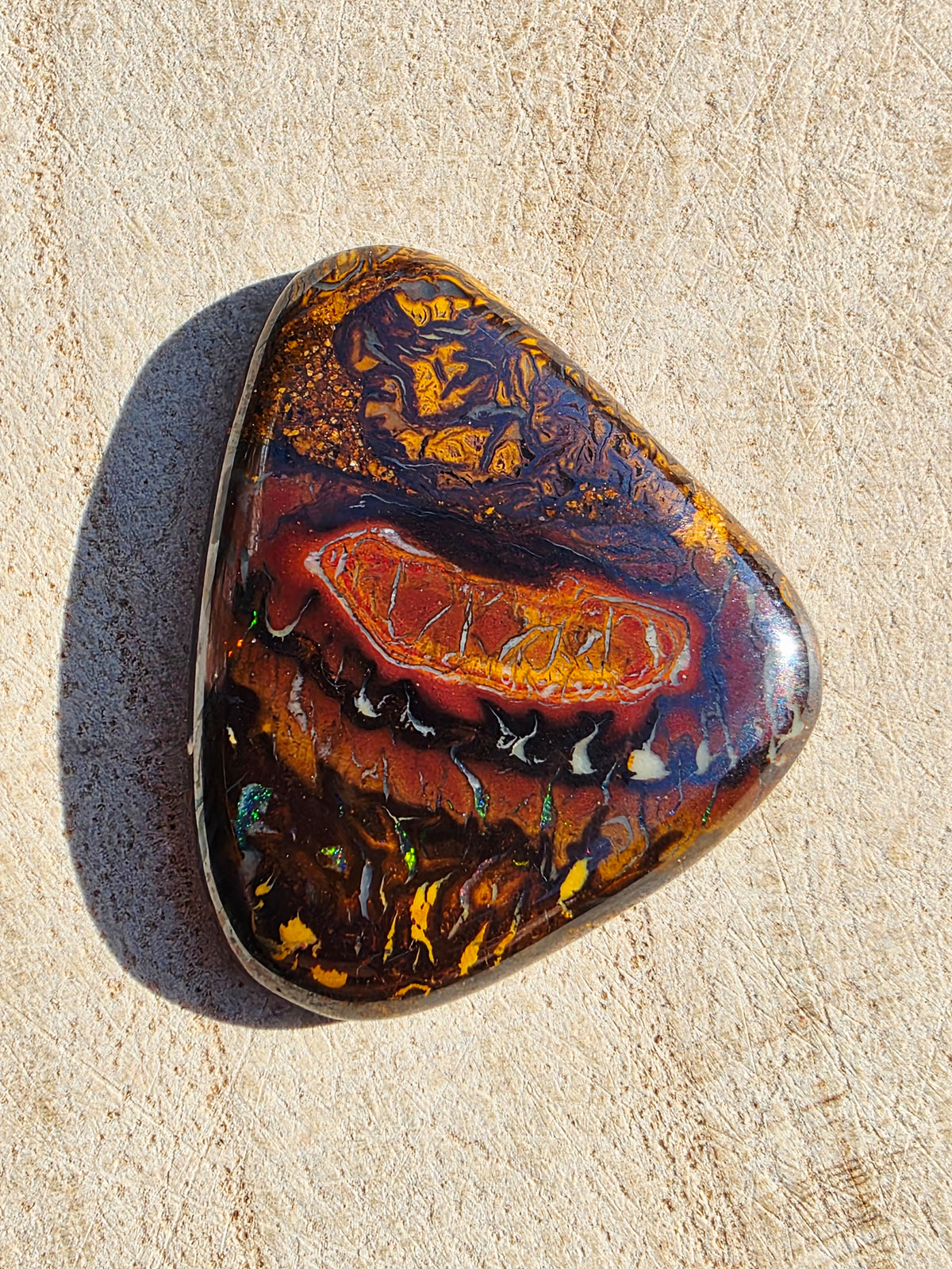 A polished, irregularly shaped boulder opal gemstone with a highly marbled pattern of gold, orange, red, brown, and black, resting on a light beige textured surface.