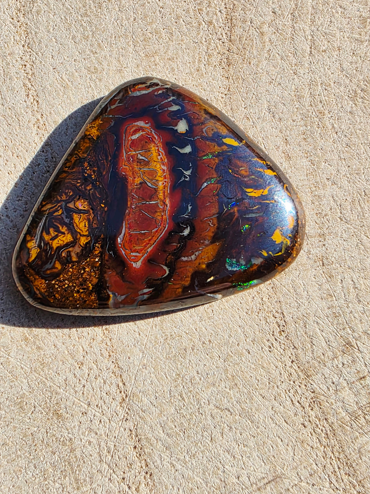 A polished triangular opal gemstone with a complex marbled pattern of orange, red, black, and gold, on a textured beige stone background.