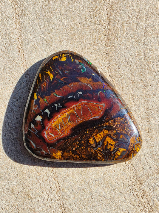 A polished triangular opal stone with a marbled pattern of black, brown, orange, and gold, resting on a textured beige surface.