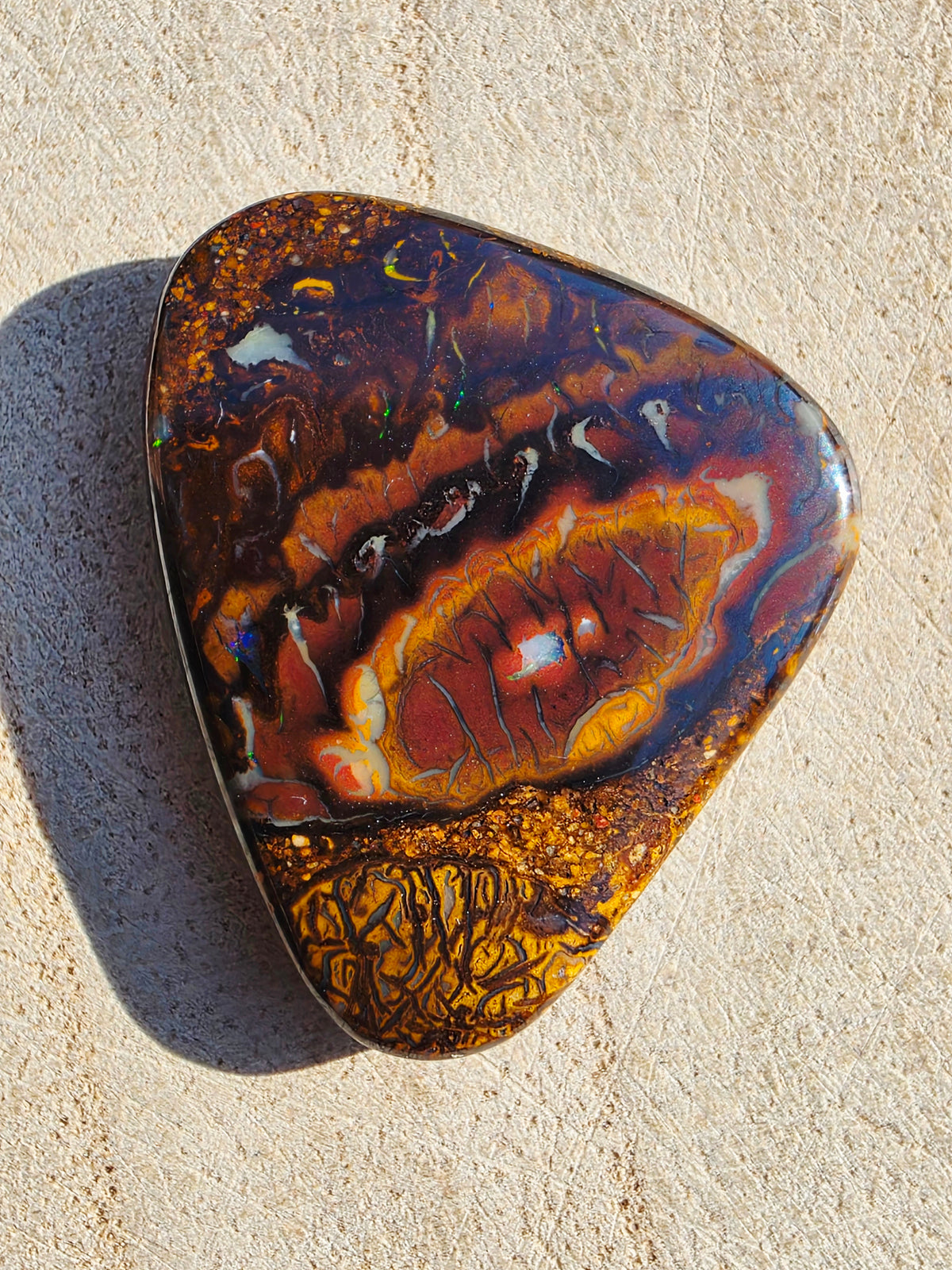 A polished boulder opal with a complex pattern of dark brown, orange, red, and gold, photographed on a textured beige surface.