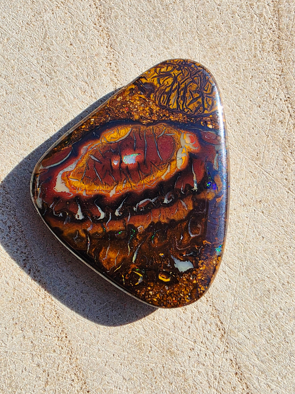 A polished natural stone opal with a complex concentric pattern of orange, red, brown, and black, resting on a textured beige stone surface.