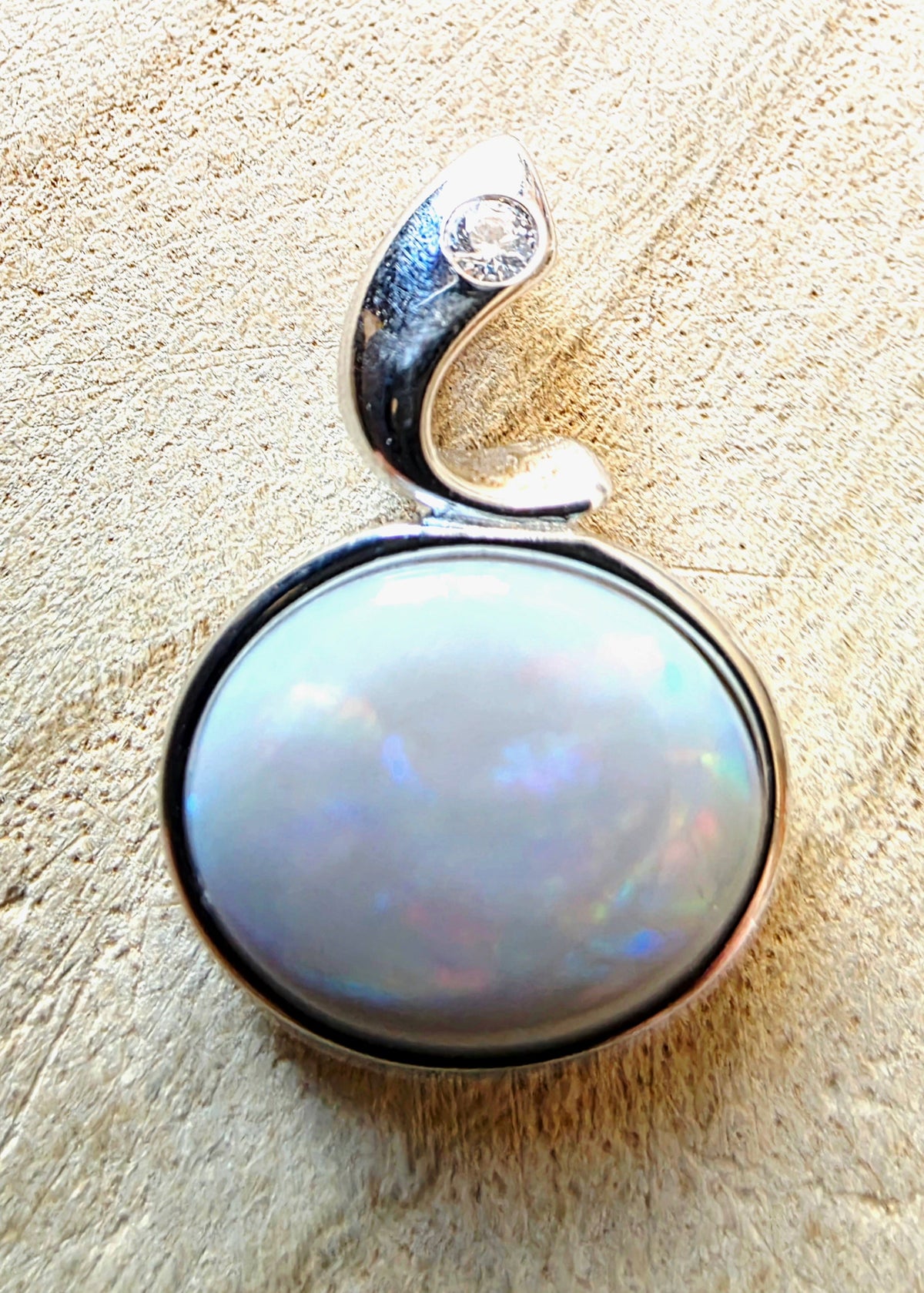 A silver pendant featuring a large, round, iridescent white opal gemstone in a bezel setting, with a curved silver bail at the top accented by a small clear stone, shown on a textured beige background.