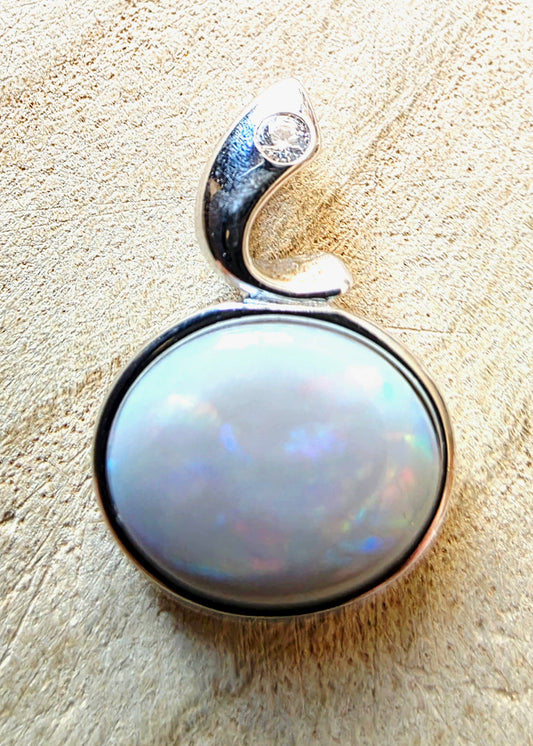A silver pendant featuring a large, round, iridescent white opal gemstone in a bezel setting, with a curved silver bail at the top accented by a small clear stone, shown on a textured beige background.