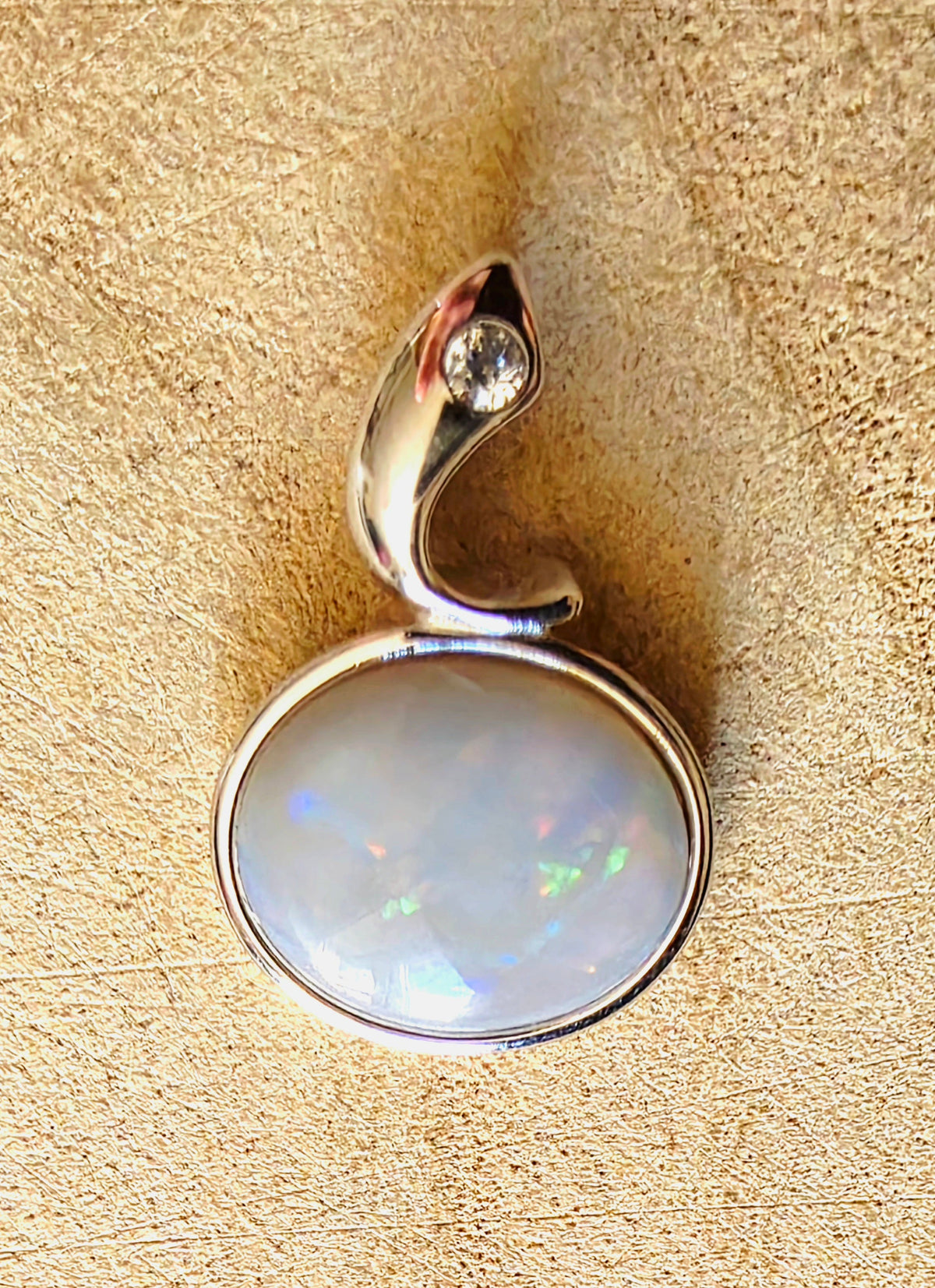 A silver pendant featuring a large oval opal cabochon opal with iridescent play-of-colour, set in a curved silver bail with a small clear accent stone, displayed on a textured beige surface.