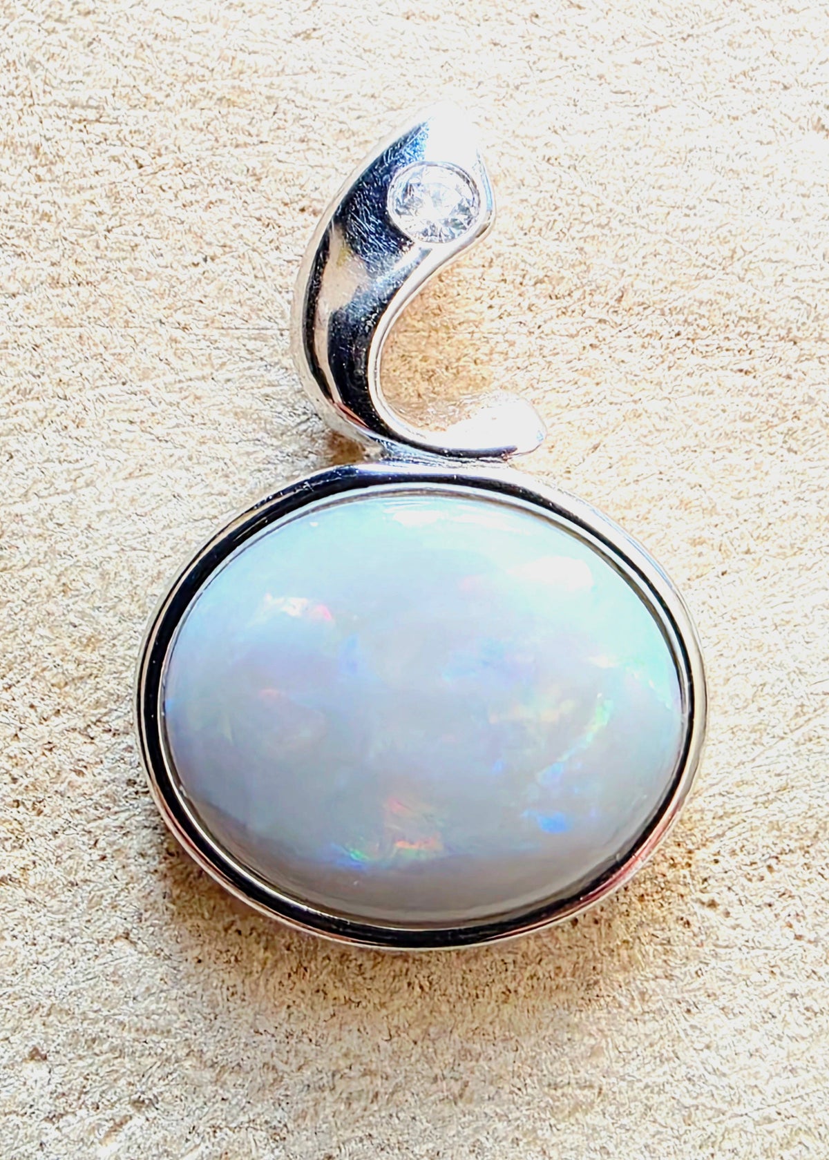 A silver pendant featuring a large, round, iridescent white opal gemstone in a polished bezel setting, with a curved silver bail on a textured beige background.
