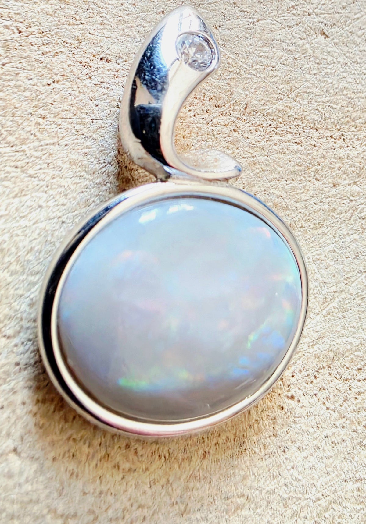 A silver pendant featuring a large, round iridescent white opal gemstone in a polished bezel setting, with a curved silver bail at the top containing a small clear accent stone, shown on a textured beige surface.