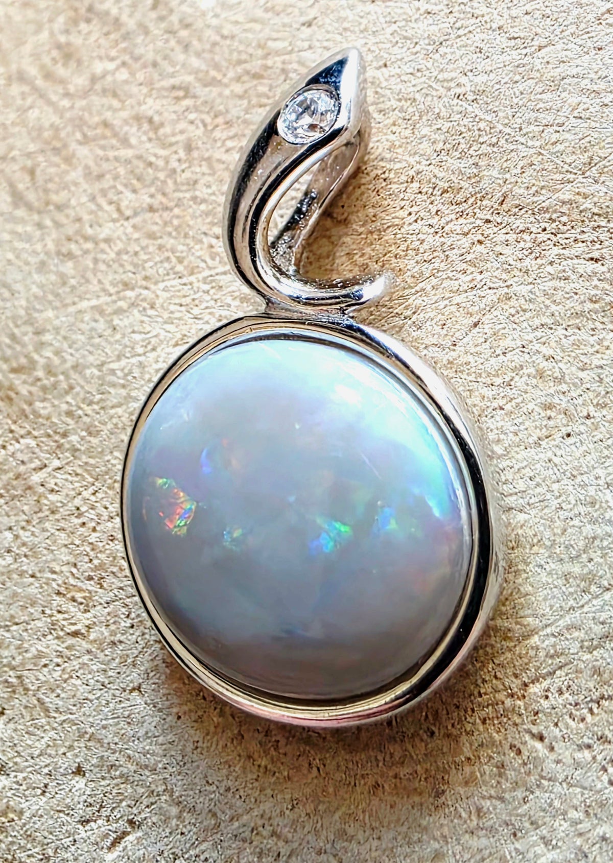 A silver pendant featuring a large, round iridescent opal gemstone with a silver bezel and a curved bail decorated with a small clear stone, shown on a textured beige surface.