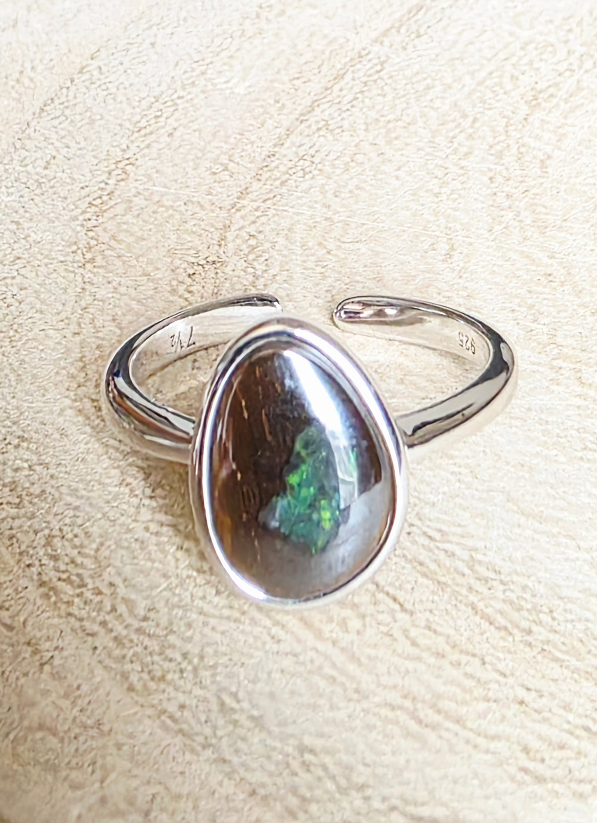 A sterling silver open band ring featuring a teardrop-shaped bezel-set brown opal with a vibrant green flash, displayed on a textured beige surface.