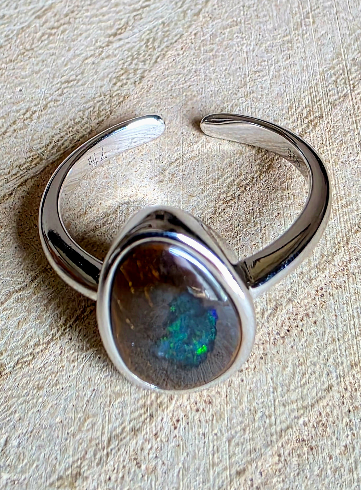 A polished silver-tone open cuff ring featuring a large teardrop-shaped cabochon stone with a dark brown base and vivid green, blue, and orange iridescent flashes, displayed on a textured beige fabric background.