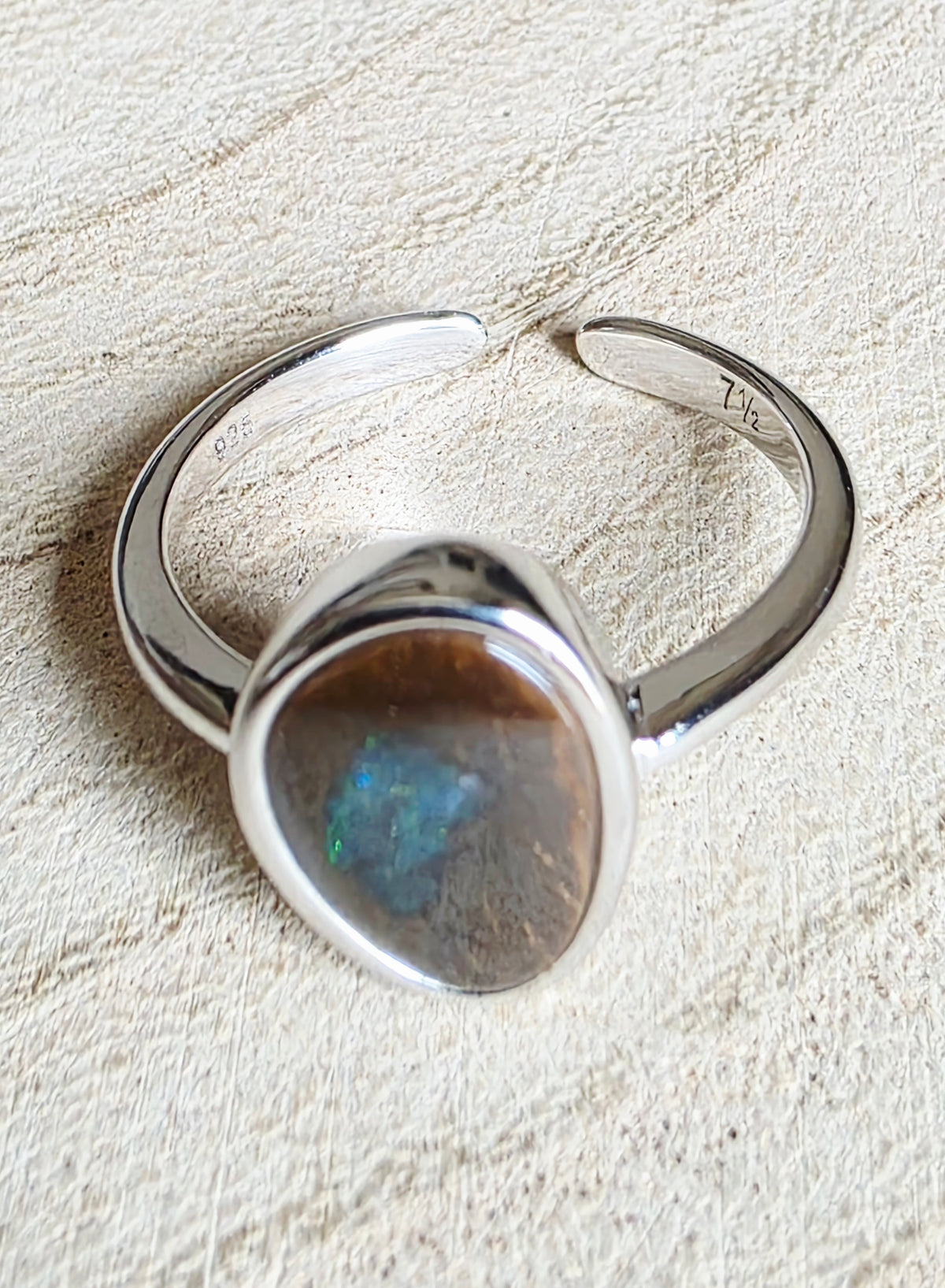 A sterling silver open band ring featuring a large, irregularly shaped cabochon stone with a brown base and a vibrant blue-green iridescent flash, displayed on a textured beige fabric background.