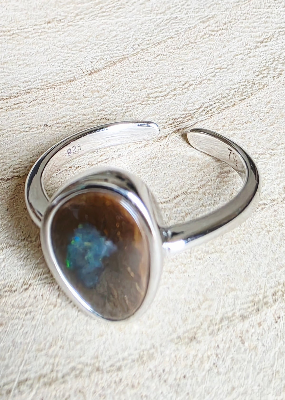A sterling silver open cuff ring featuring a large, teardrop-shaped opal stone with iridescent blue flashes, resting on a textured beige fabric background.