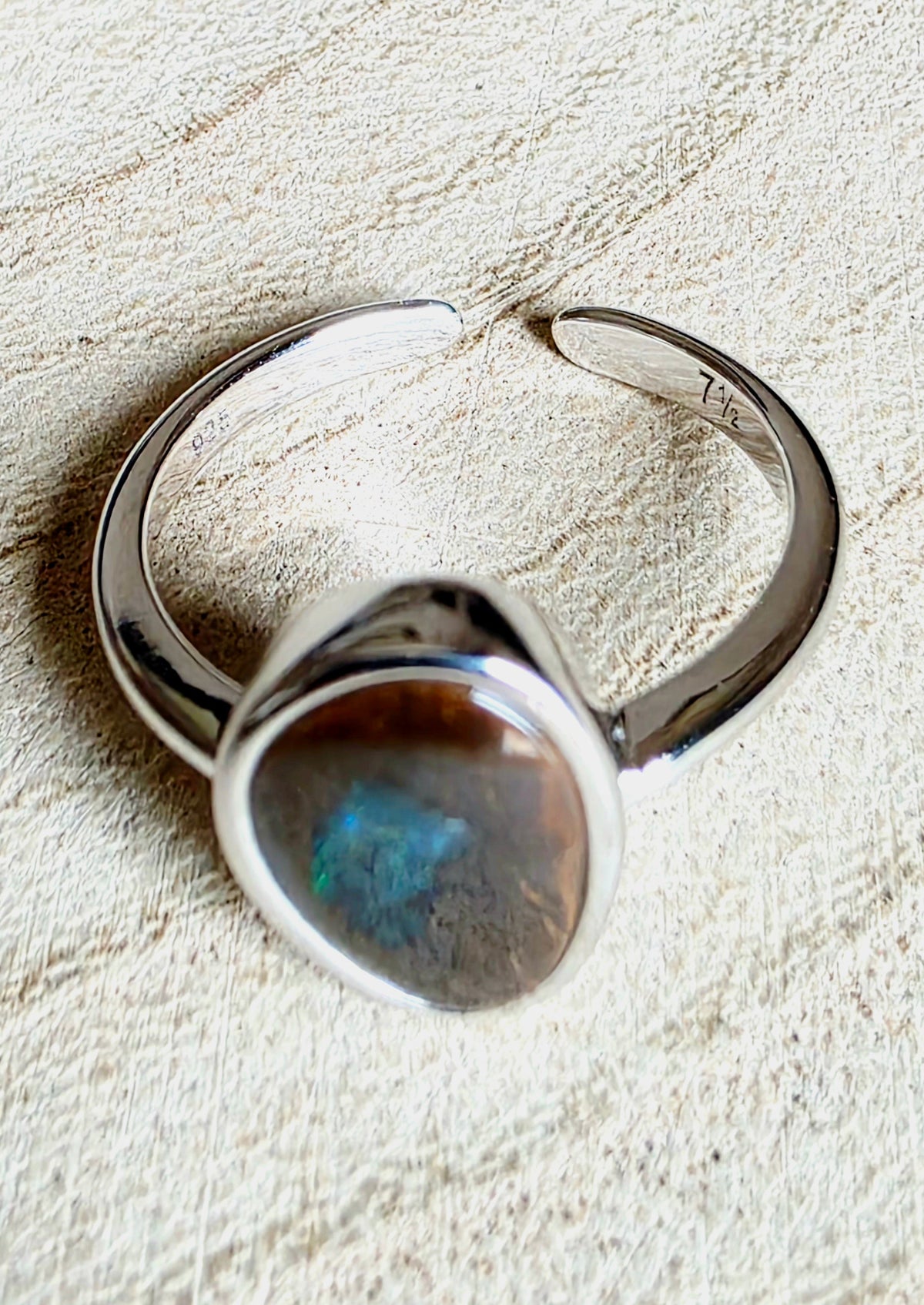 A sterling silver open band ring featuring a large, polished oval cabochon stone with a brown and blue iridescent play-of-colour, displayed on a textured beige fabric background.