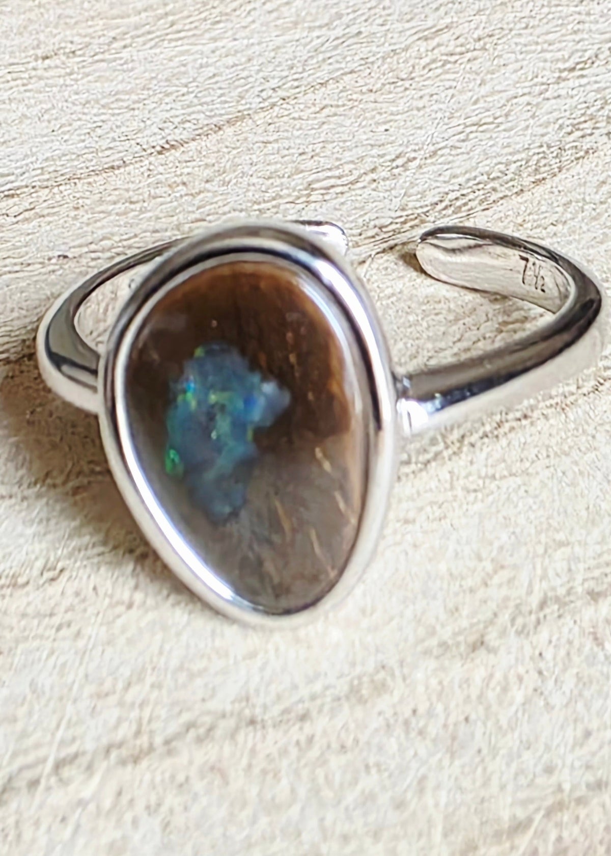 A silver open band ring featuring a large, irregularly shaped brown opal stone with vibrant blue and green flashes, resting on a textured beige surface.