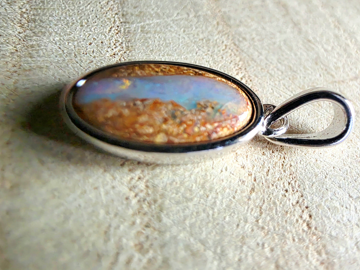 An oval pendant featuring a colourful opal cabochon with orange, blue, and gold tones, set in a polished silver bezel with a hanging loop, resting on a textured beige surface.