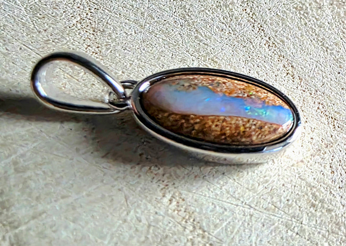 A silver oval pendant featuring a brown opal stone with vibrant blue and green iridescent play-of-colour, set in a polished silver bezel with a hanging loop.