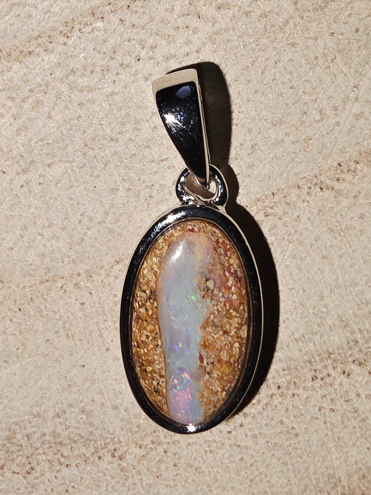 An oval pendant featuring a natural opal stone with iridescent colours set in a silver bezel, displayed on a textured beige surface.