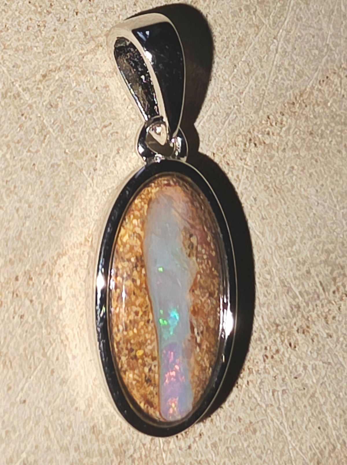 An oval pendant featuring a natural opal stone with iridescent colours set in a polished silver bezel, shown on a textured beige background.