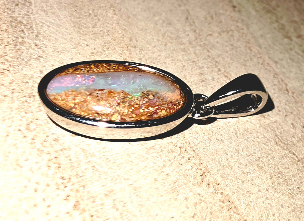 An oval pendant with a silver border and bail, featuring a colourful inlay of gold flakes and iridescent blue-green opal-like material, resting on a textured beige surface.