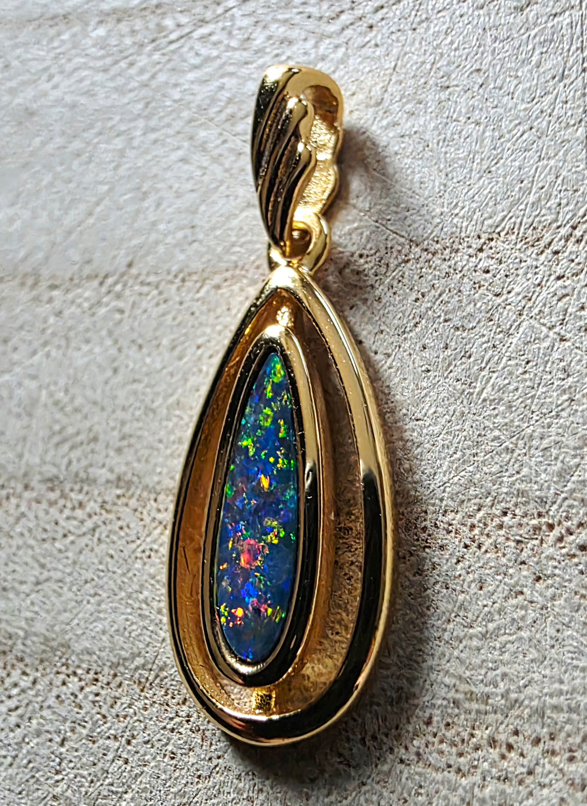 A gold teardrop pendant featuring a vibrant, multicoloured opal inlay, photographed close-up on a textured light beige surface.