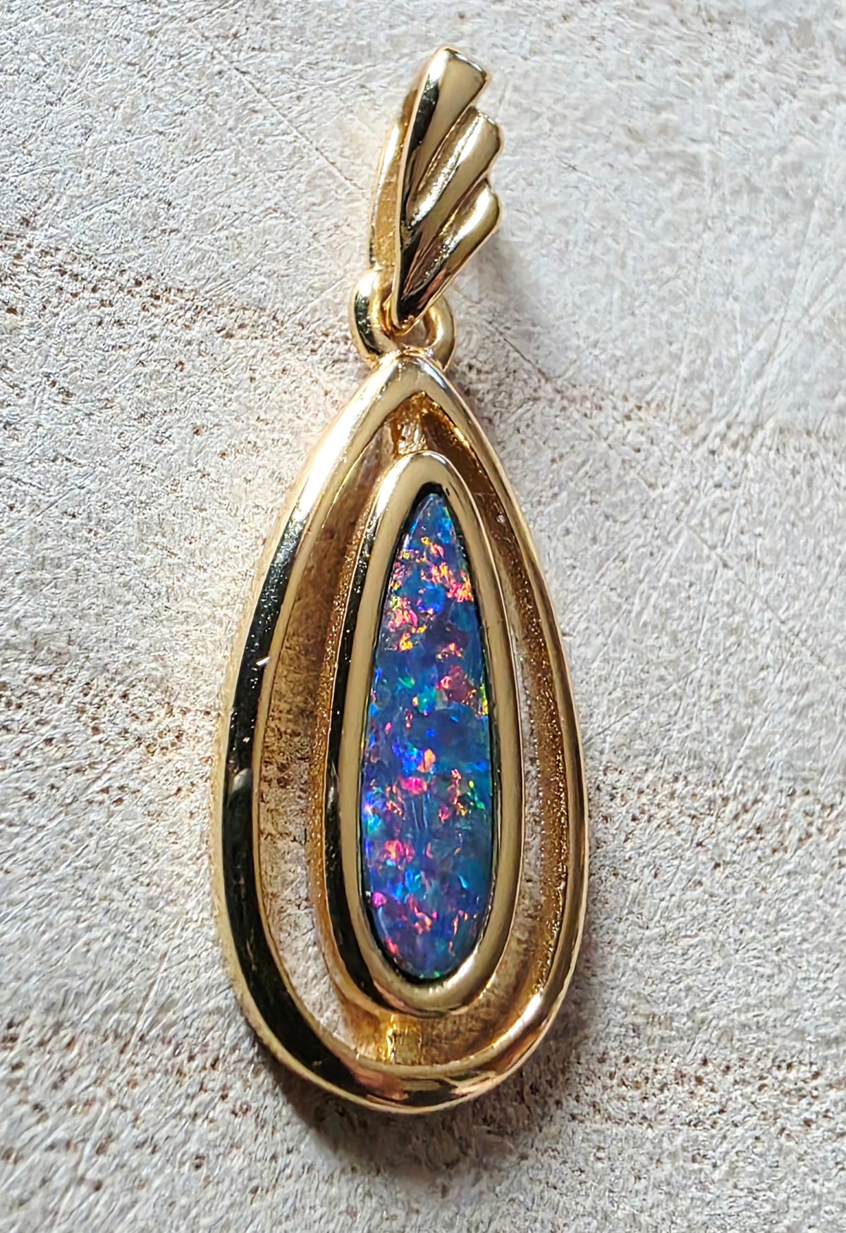 A gold teardrop pendant featuring a central iridescent opal stone with blue, pink, and green flashes, set against a textured light beige background.
