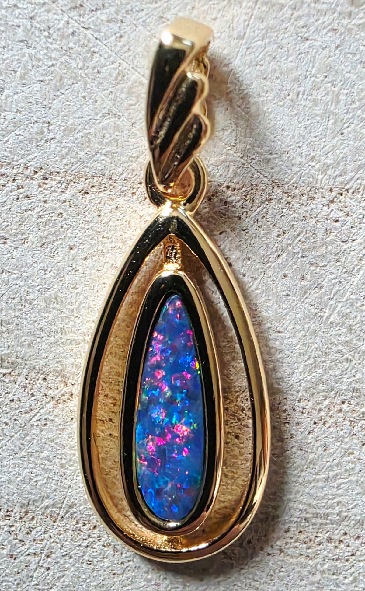 A gold teardrop pendant featuring a vibrant blue and pink red iridescent opal inlay, set against a textured light beige background.