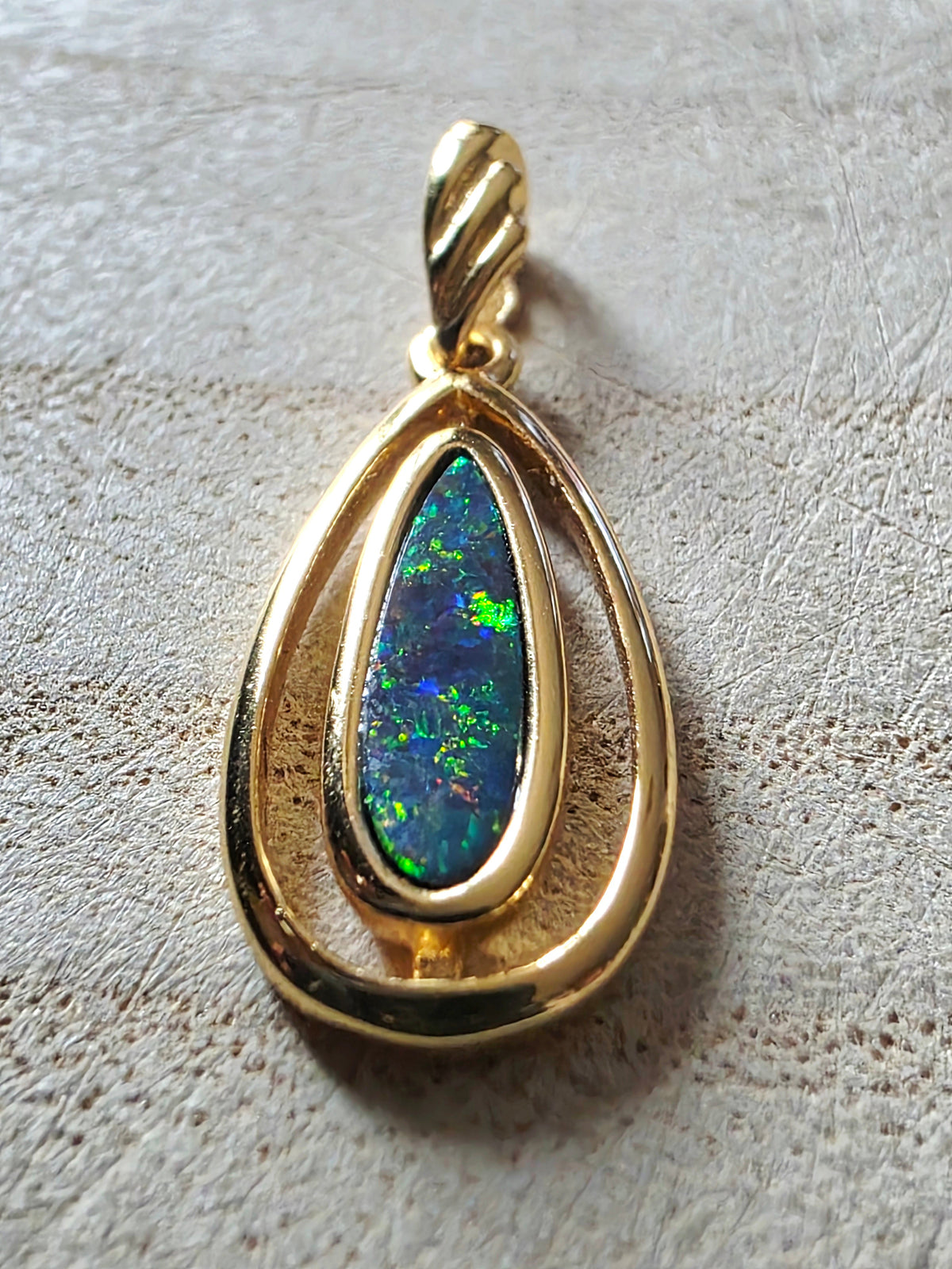 A gold teardrop pendant featuring a central teardrop-shaped iridescent opal stone with green, blue, and orange flashes, set in a polished gold frame with a twisted bail at the top, resting on a textured beige surface.