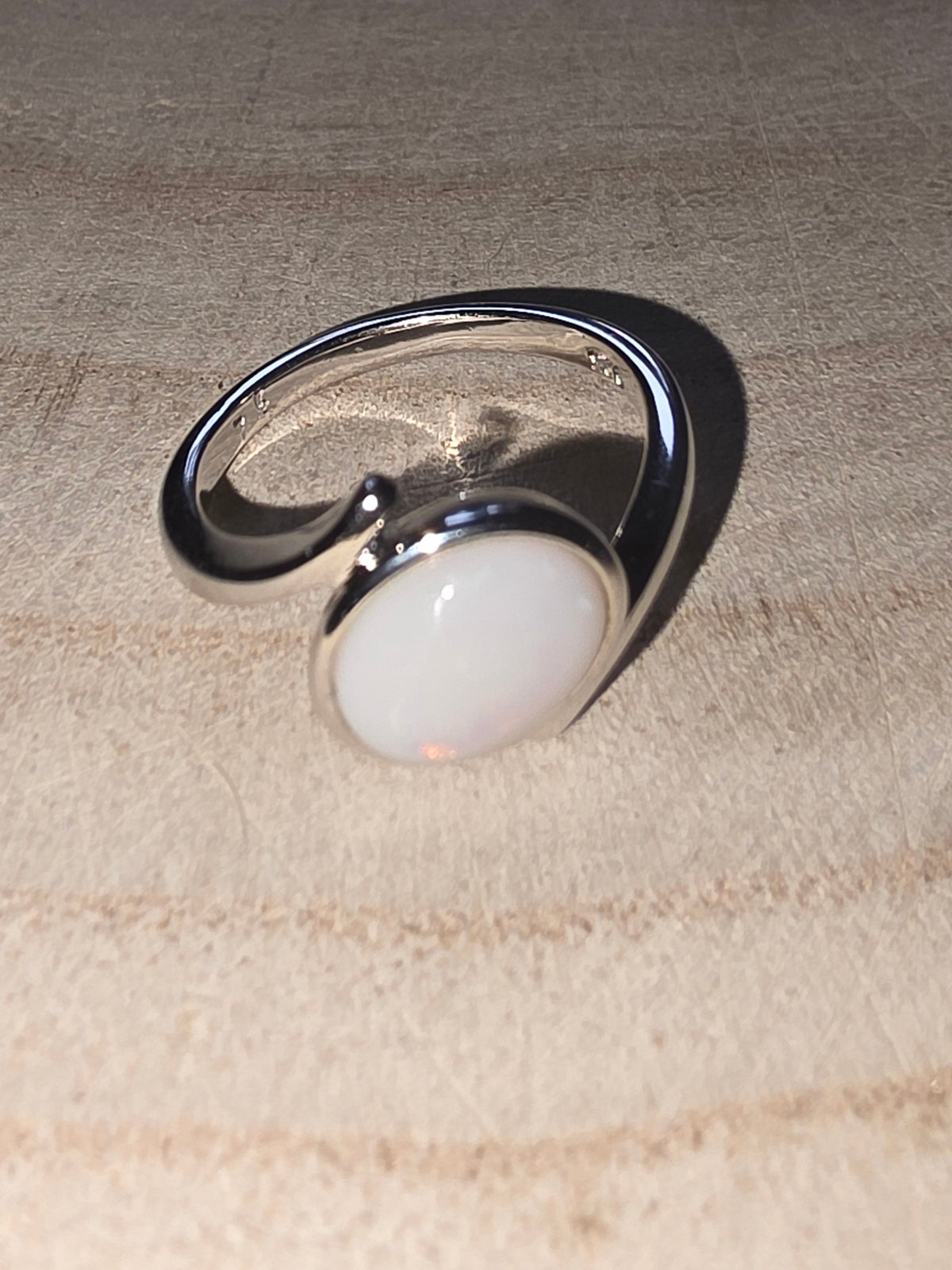 A silver open band ring featuring a large, smooth, oval crystal opal cabochon stone, displayed on a textured beige surface.