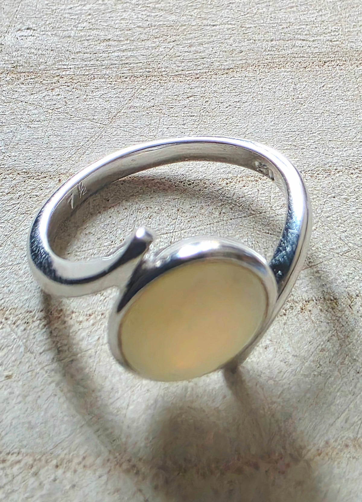 A sterling silver open-band ring with a large, smooth, oval-shaped pale cream or yellowish-beige opal, resting on a textured light beige surface.