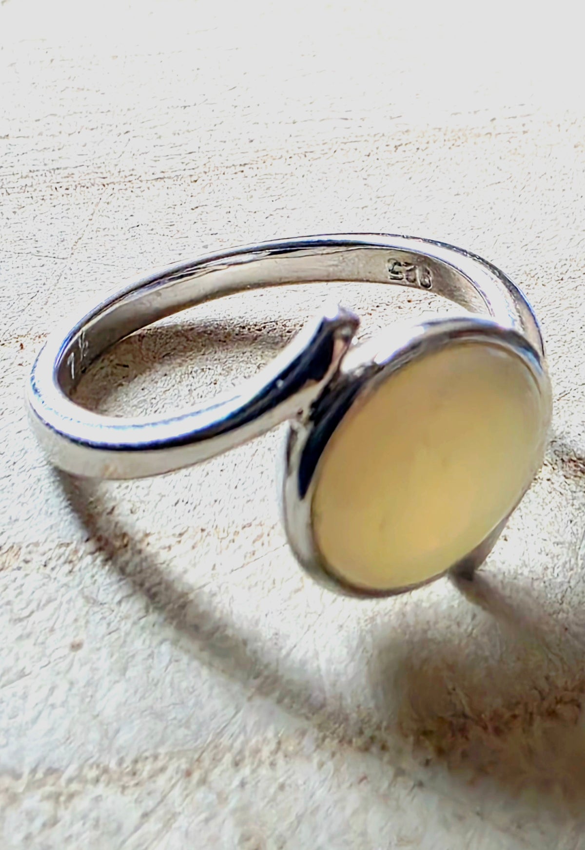 A sterling silver open-band ring with a large, smooth, oval pale yellow stone, photographed on a textured beige surface.