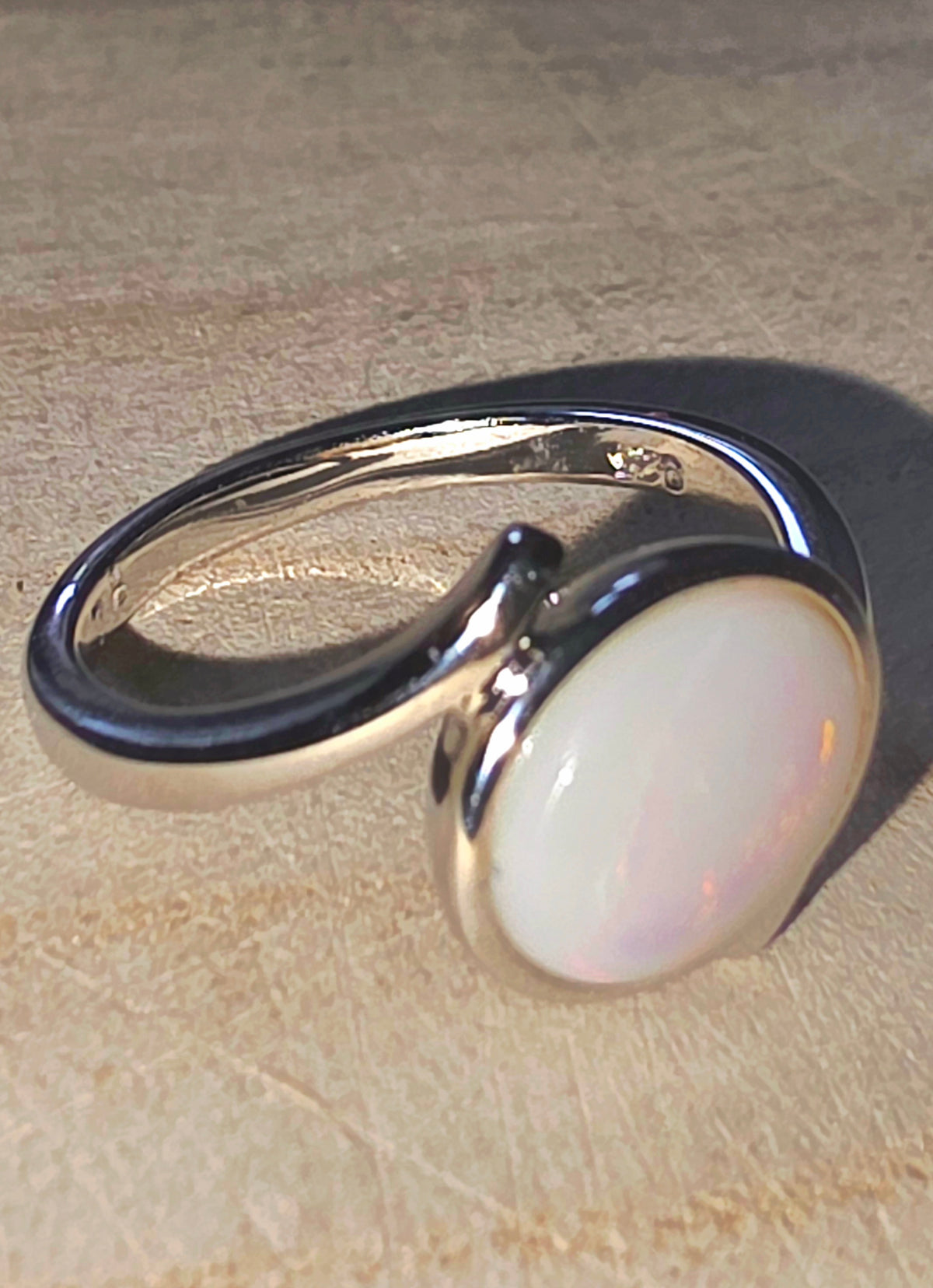 A silver-toned ring with a large, smooth, oval white cabochon opal, photographed on a textured beige surface.