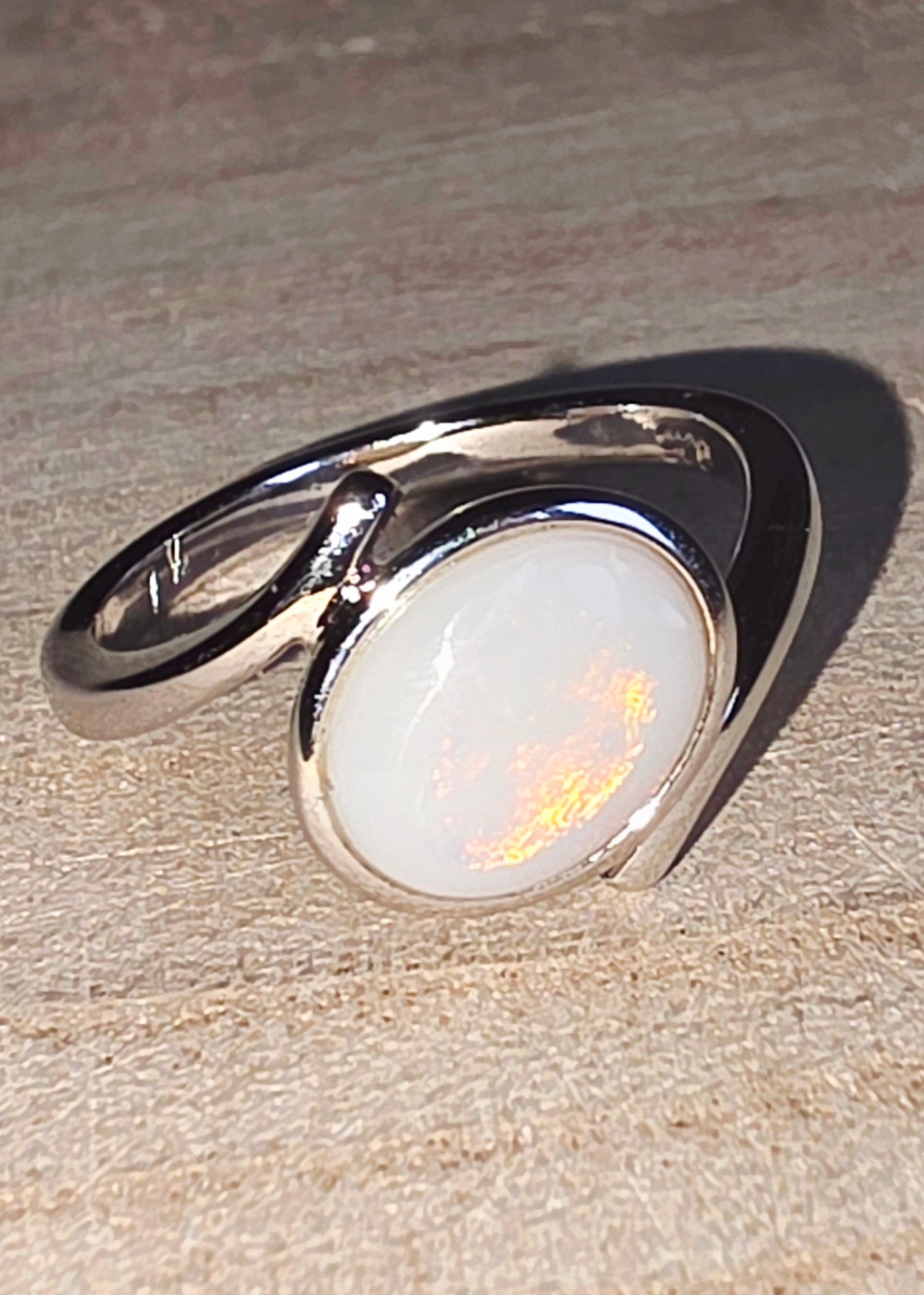 A silver ring featuring a large, oval-shaped crystal opal stone with a warm orange-gold iridescent flash, resting on a textured beige surface.