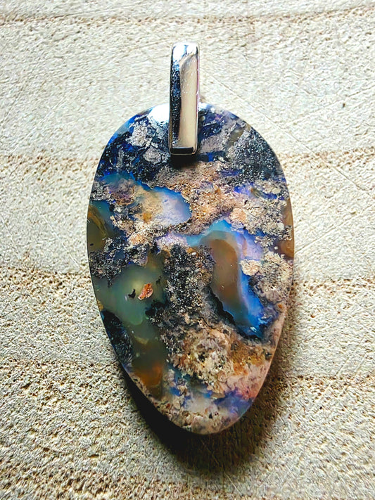 A teardrop-shaped pendant featuring a natural opal with vibrant blue, brown, black, and cream patterns, suspended from a polished silver 925 bail, set against a textured beige background.