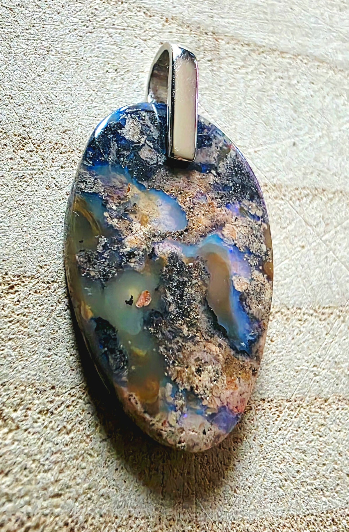 An oval pendant featuring a highly iridescent, multi-coloured opal with blue, gold, brown, and green tones, set in a polished silver 925 bail, displayed on a textured beige background.