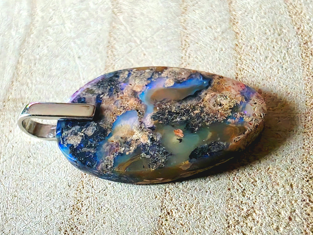 An oval pendant featuring a highly marbled, opalescent opal with blue, gold, brown, and black patterns, attached to a polished silver 925 bail, resting on a textured beige background.