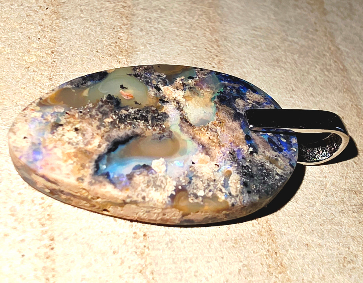A polished oval pendant featuring a natural iridescent opal with blue, purple, cream, and brown tones, displayed on a sandy beige surface.