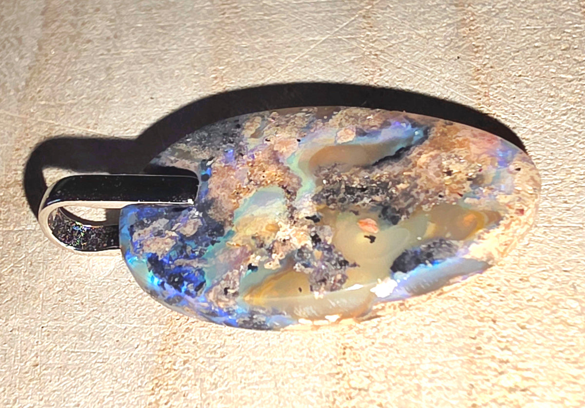 An oval-shaped opal pendant with a black metal bail, resting on a textured beige surface. The opal features a complex play-of-colour pattern with blue, green, yellow, and brown tones.
