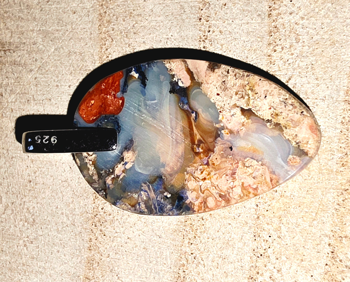 A teardrop-shaped polished opal stone pendant with swirling patterns of blue, grey, orange, and beige, displayed on a textured beige surface.