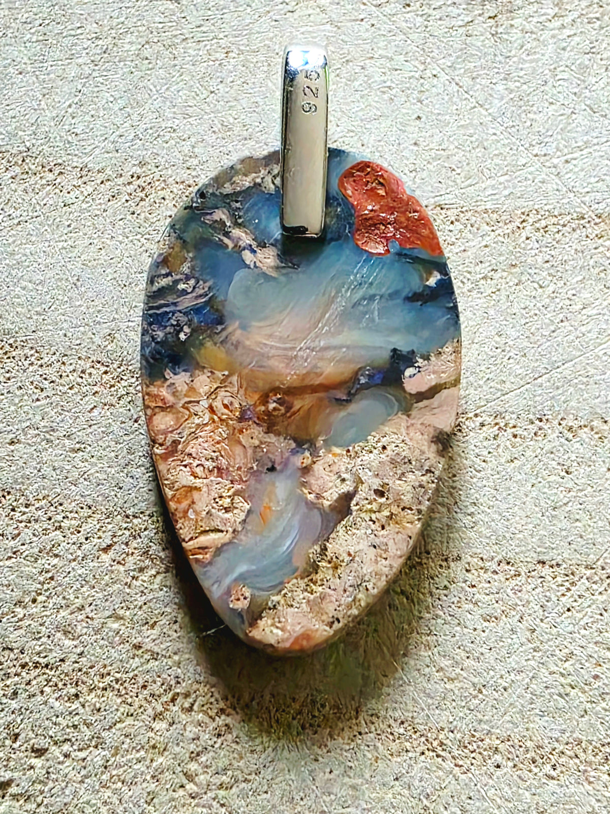 A teardrop-shaped pendant made from polished opal with swirling patterns of blue, beige, brown, and rust orange, featuring a silver bail